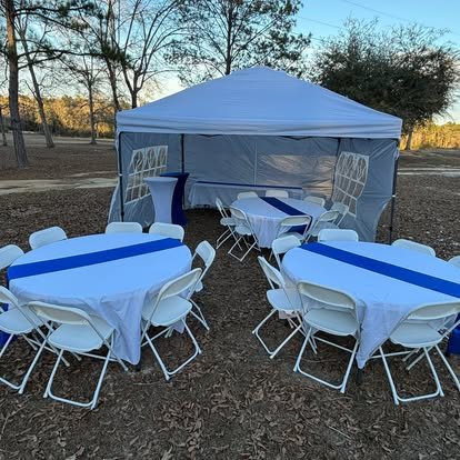 Outdoor event setup with round tables covered with white tablecloths and blue runners, white folding chairs, and a white canopy tent in a park-like setting with trees.
