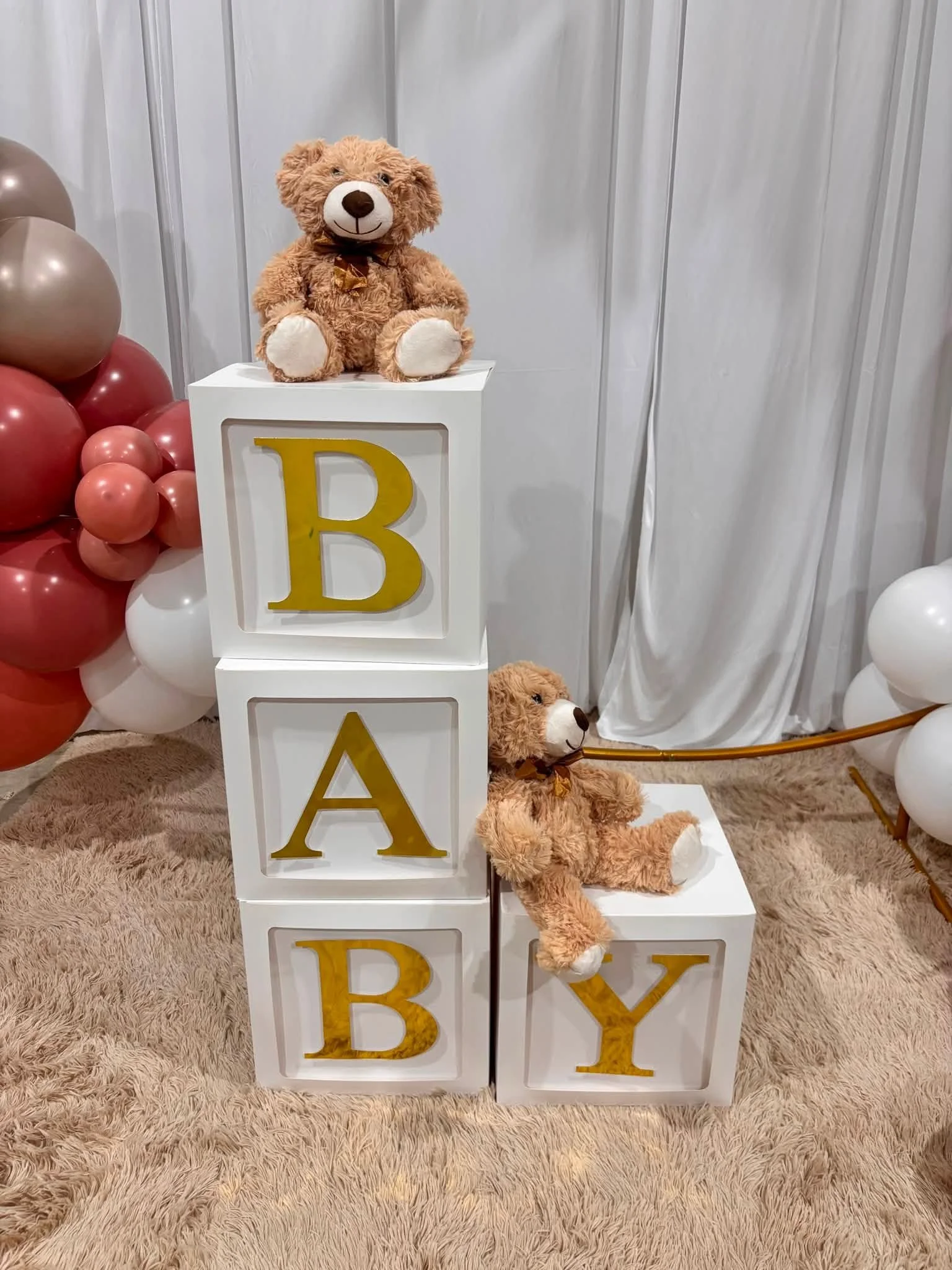 Decorative display featuring a plush teddy bear with a brown bow tie, sitting on top of white storage blocks with gold letter decals spelling 'B', 'A', 'B', 'Y'. Balloons in shades of gold, pink, red, and white surround the blocks, set on a plush beige carpet with a light grey striped wall and white curtain backdrop.