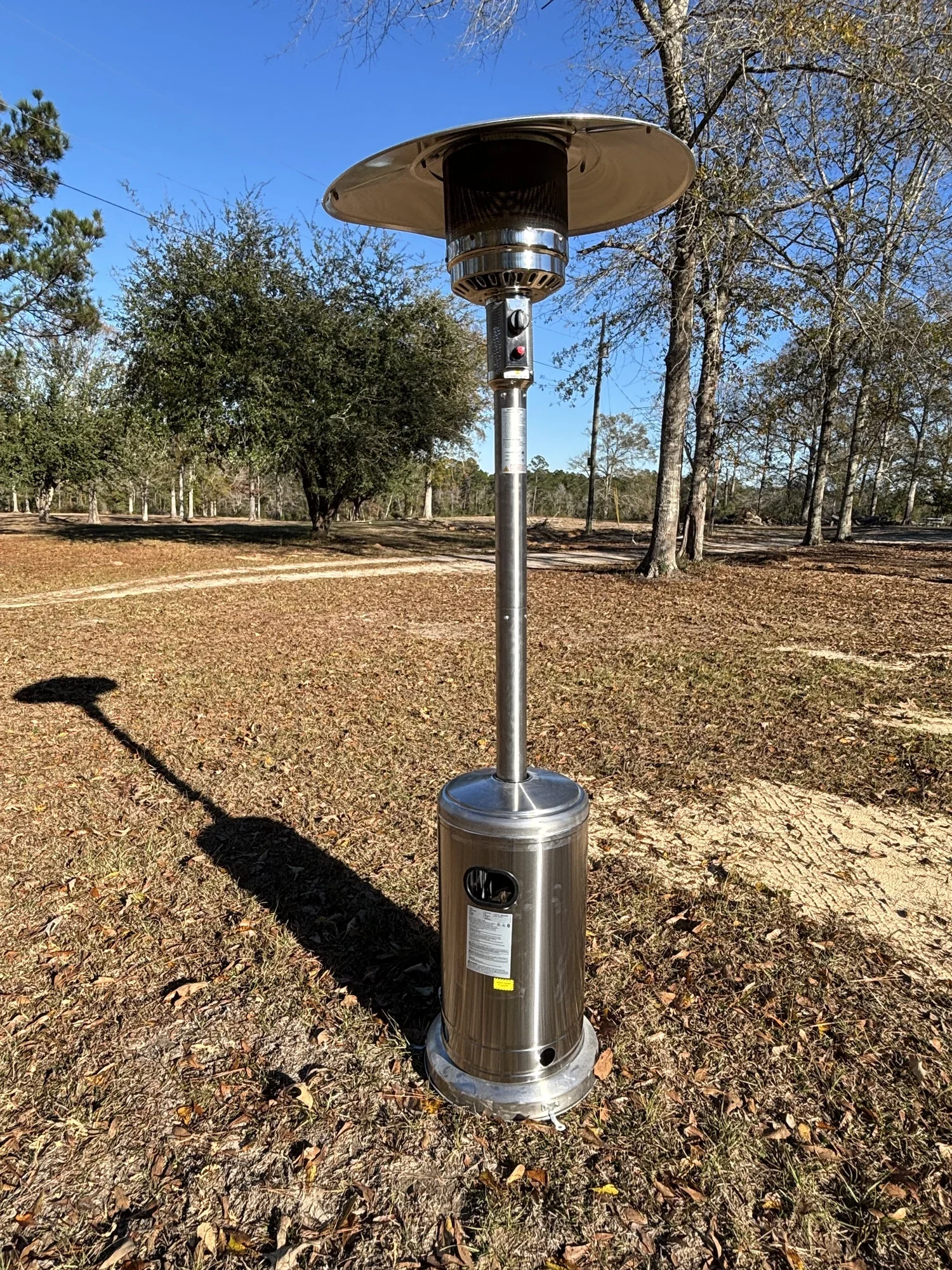 Outdoor space with a stainless steel patio heater on a leaf-covered ground, with trees and a clear blue sky in the background.