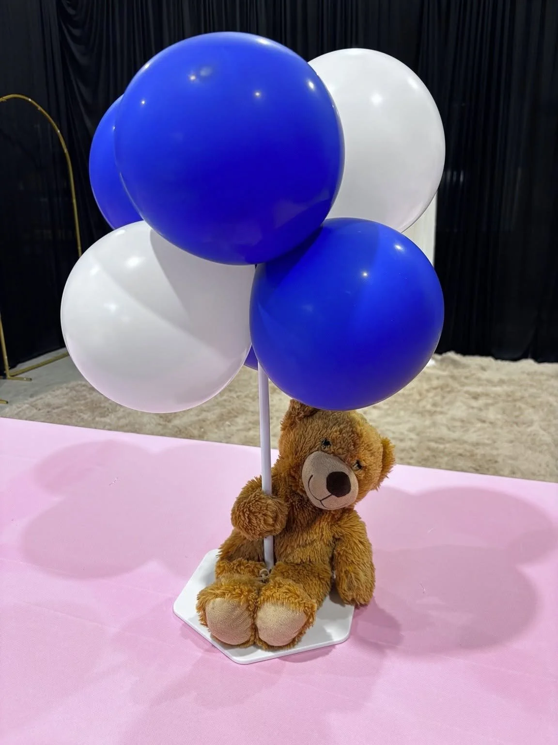 A plush teddy bear holding a white pole with blue and white balloons attached, all placed on a pink surface with a black curtain background.
