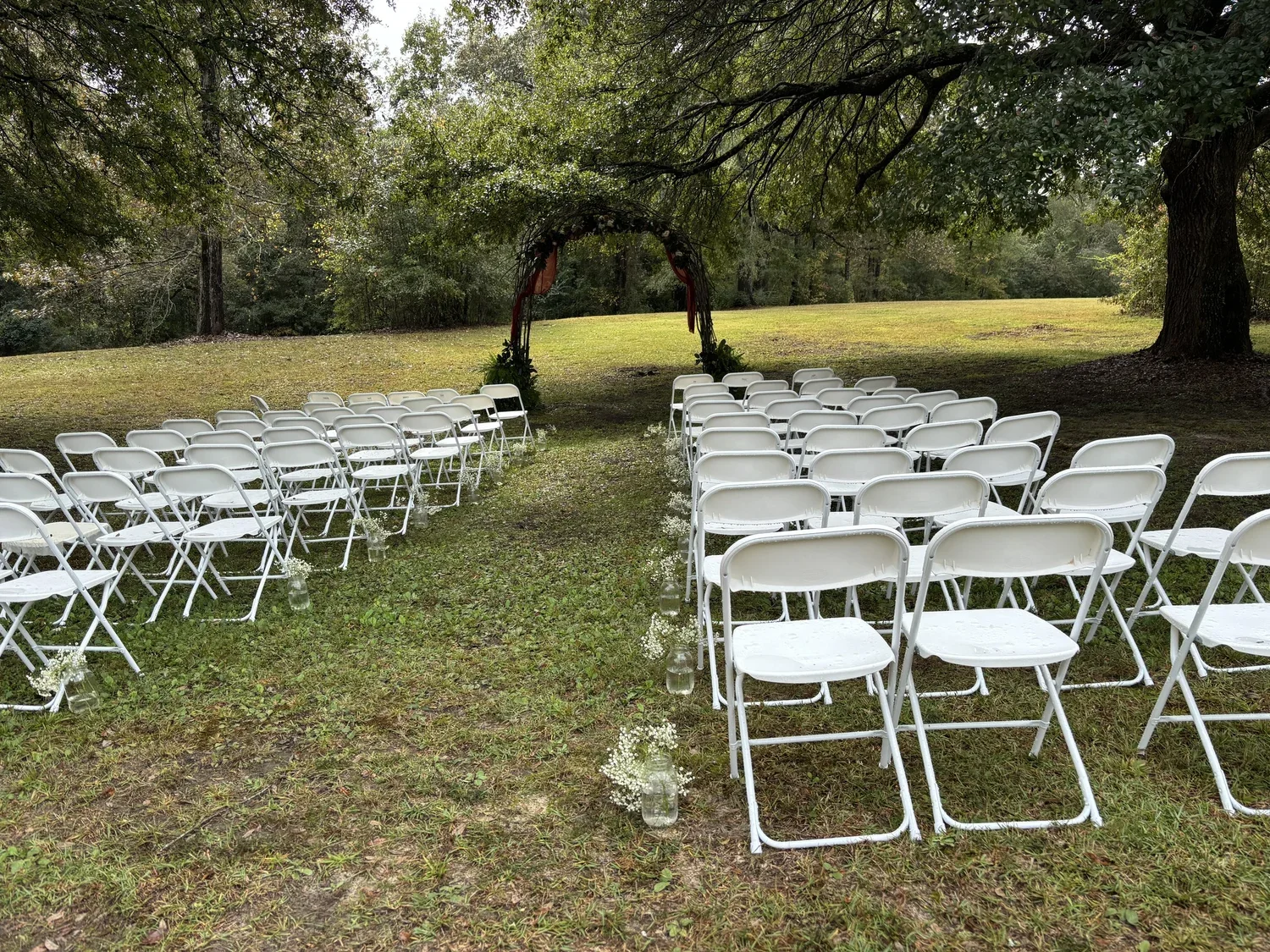 Outdoor wedding ceremony setup with white folding chairs arranged in rows and small floral decorations in jars along the aisle, under a wooden arch with draped fabric, in a grassy area surrounded by trees.