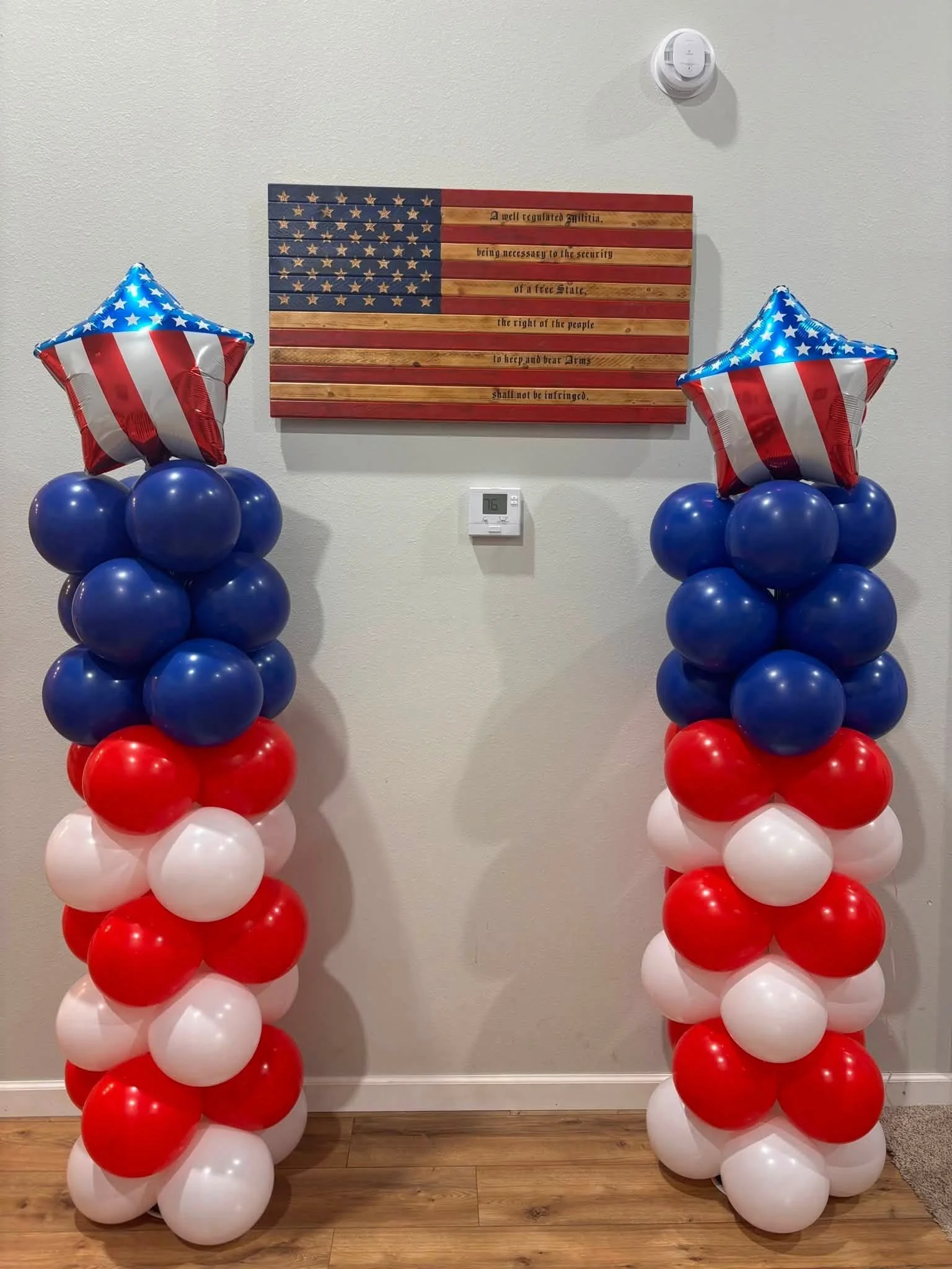 Two balloon columns with red, white, and blue balloons topped with star-shaped balloons resembling the American flag, positioned on either side of a wooden American flag sign with an engraved quote.