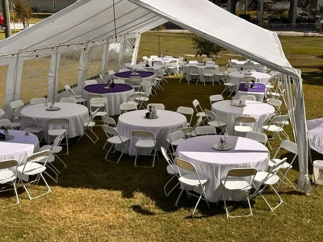 Outdoor event tent with round tables covered in white tablecloths, purple table runners, and white chairs on grass, set up for a gathering or celebration.