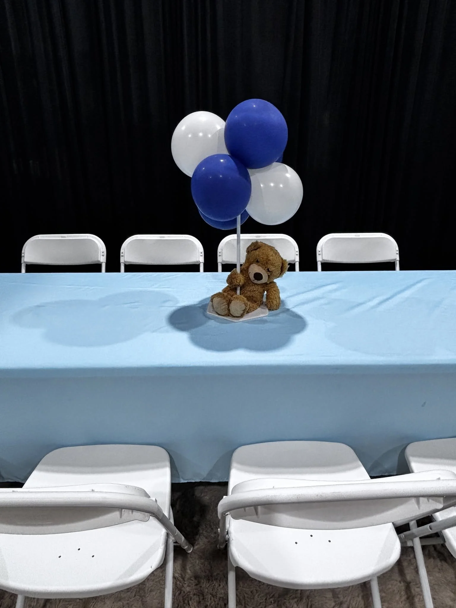 A table with a blue tablecloth and a centerpiece of five balloons (white and blue) and a teddy bear holding the balloon stick, set against a backdrop of black curtains with white chairs arranged on either side.