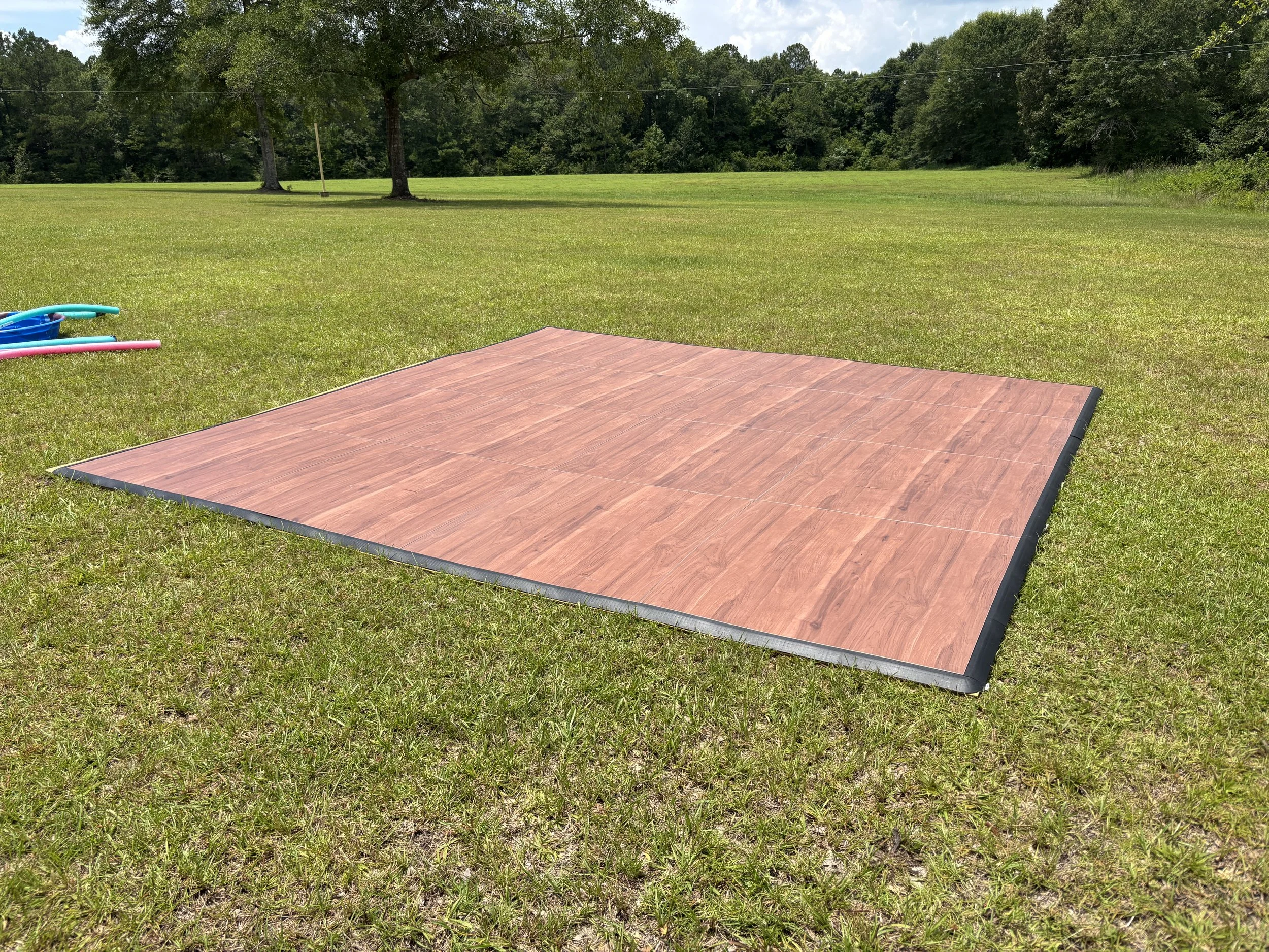 A backyard scene with a large grassy area, a couple of trees, and a wooden platform on the grass. There are some plastic pool toys in the corner and background features a forested area with a bright, partly cloudy sky.