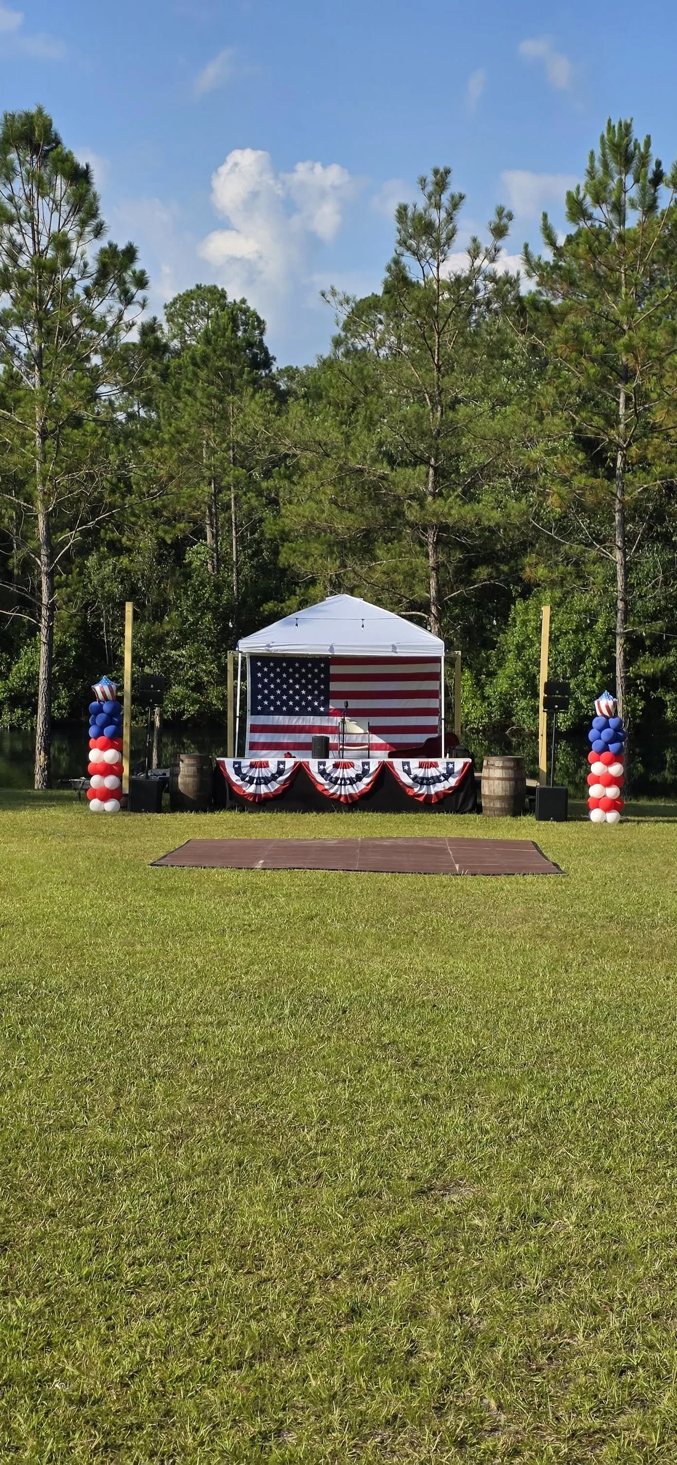 Outdoor celebration setup with a stage decorated with American flags, patriotic bunting, and balloon columns, surrounded by green grass and tall trees under a partly cloudy blue sky.