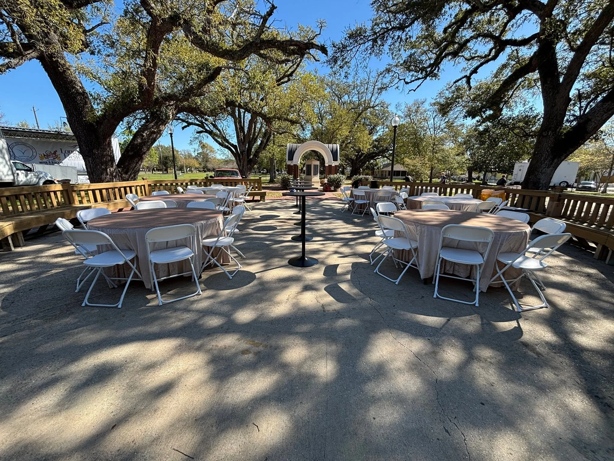 Outdoor event space with round tables covered in beige tablecloths and white folding chairs, surrounded by large trees with sprawling branches, and pathways, in a park-like setting on a sunny day.