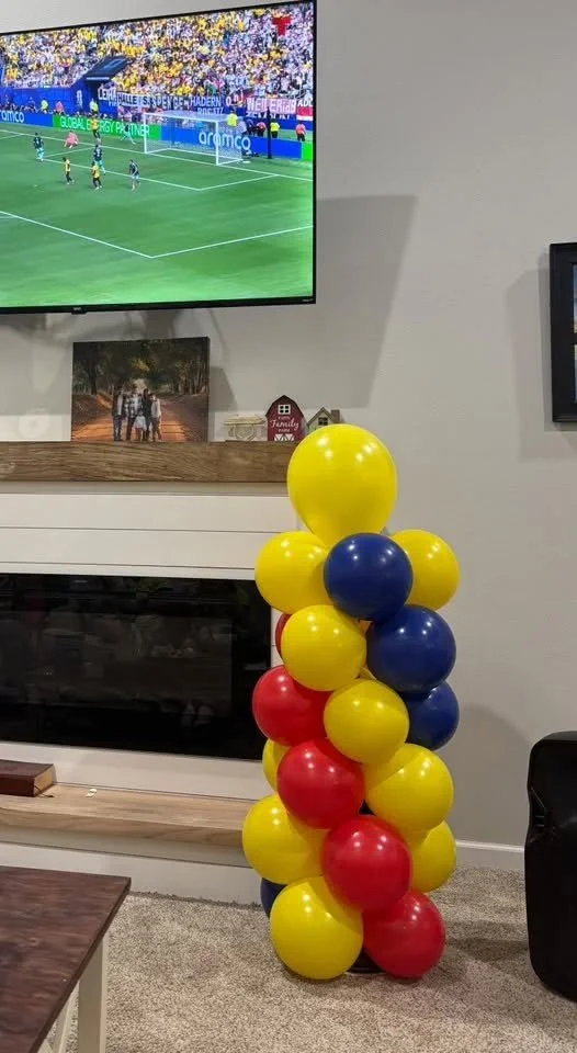 A cluster of yellow, blue, and red balloons stacked next to a fireplace in a living room, with a flat-screen TV showing a soccer game mounted on the wall above.