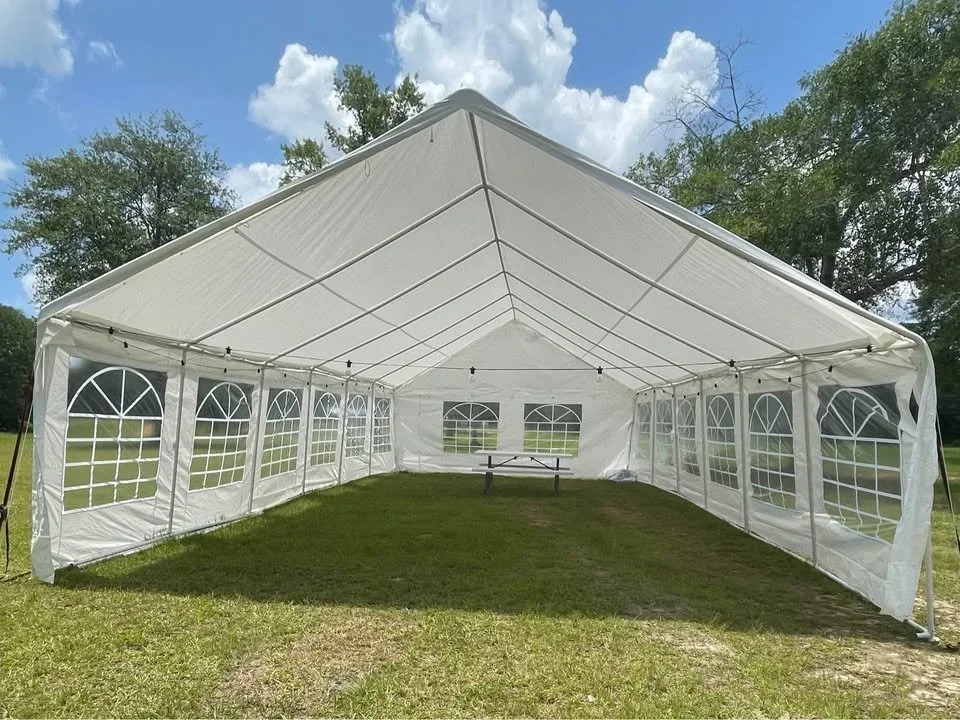 A large white event tent set up outdoors on a grassy area, with windows along the sides and a picnic table inside, under a partly cloudy sky.