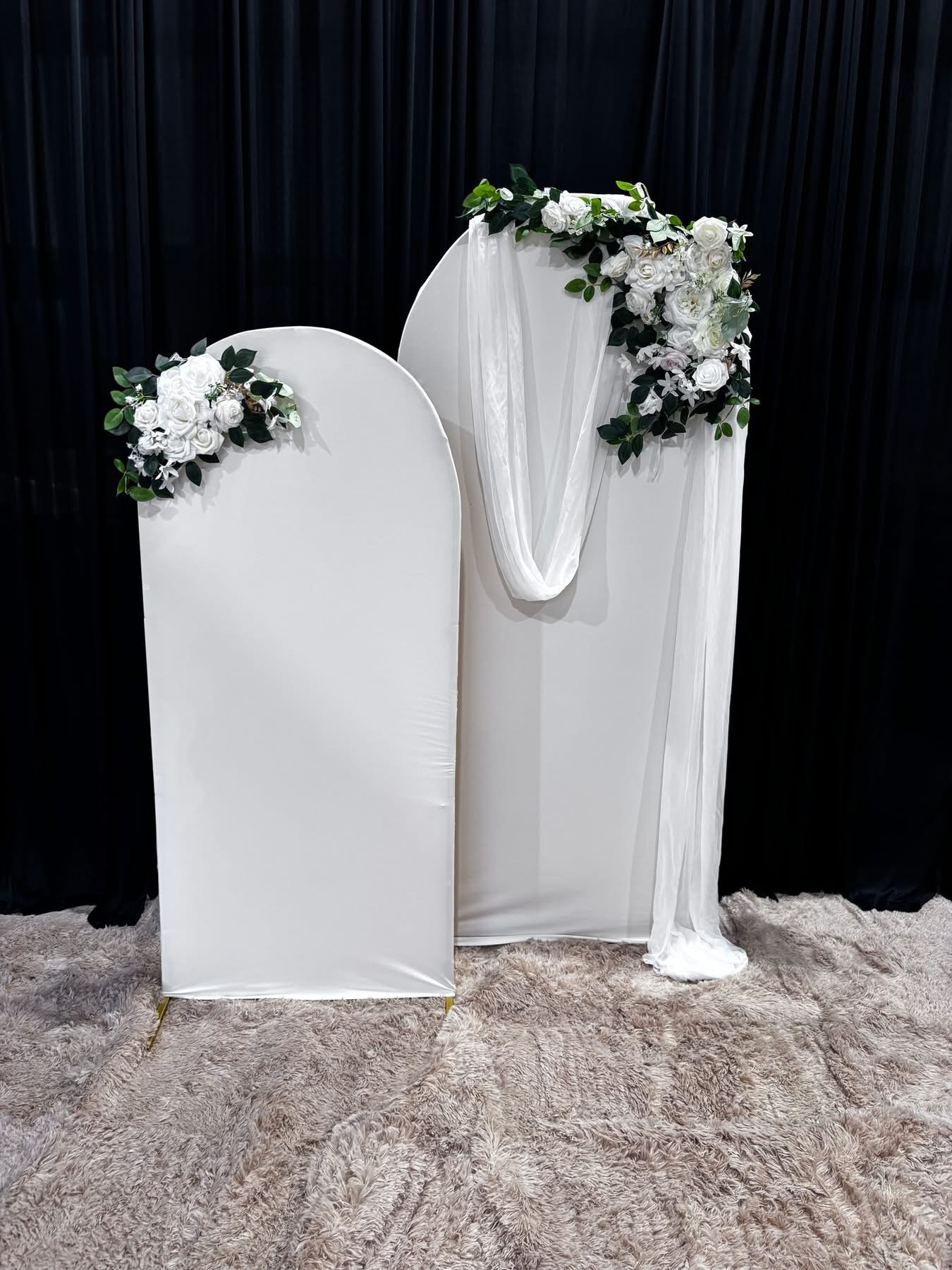 A decorative white backdrop with floral arrangements in the top corners, set on a beige shaggy carpet with a black curtain in the background.