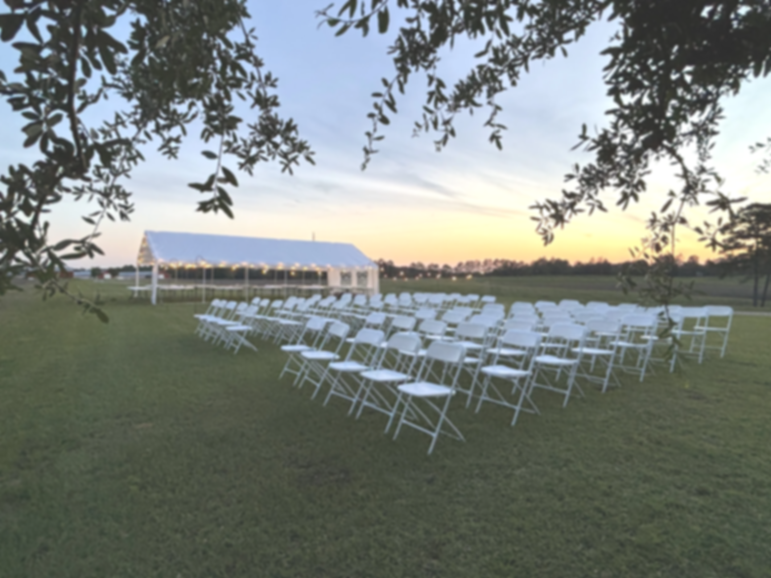 An outdoor wedding ceremony area at sunset with white chairs arranged in rows on grass, a small pavilion with a metal roof, and a body of water in the background, framed by overhanging tree branches.