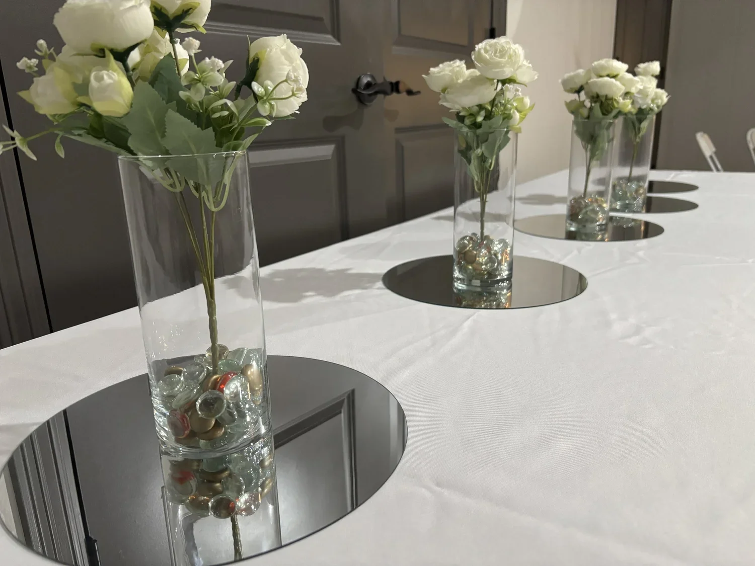 Three tall glass vases with white flowers and decorated with colorful marbles inside, placed on mirrors on a white tablecloth. A dark door is in the background.
