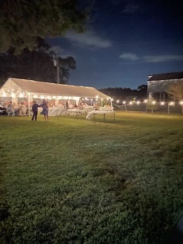 Outdoor evening event with a large illuminated tent, tables, and chairs on a grassy field, with string lights and a building in the background.
