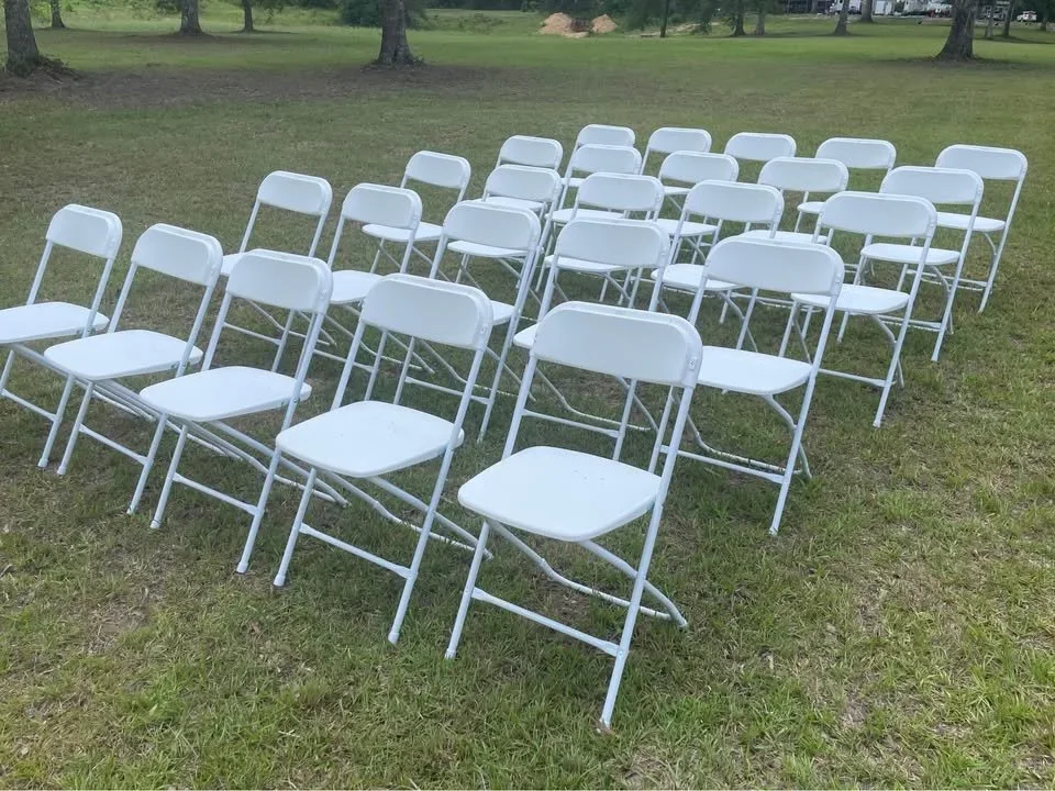 Multiple white folding chairs arranged outdoors on grass in a park or open field, with trees and a few buildings in the background.