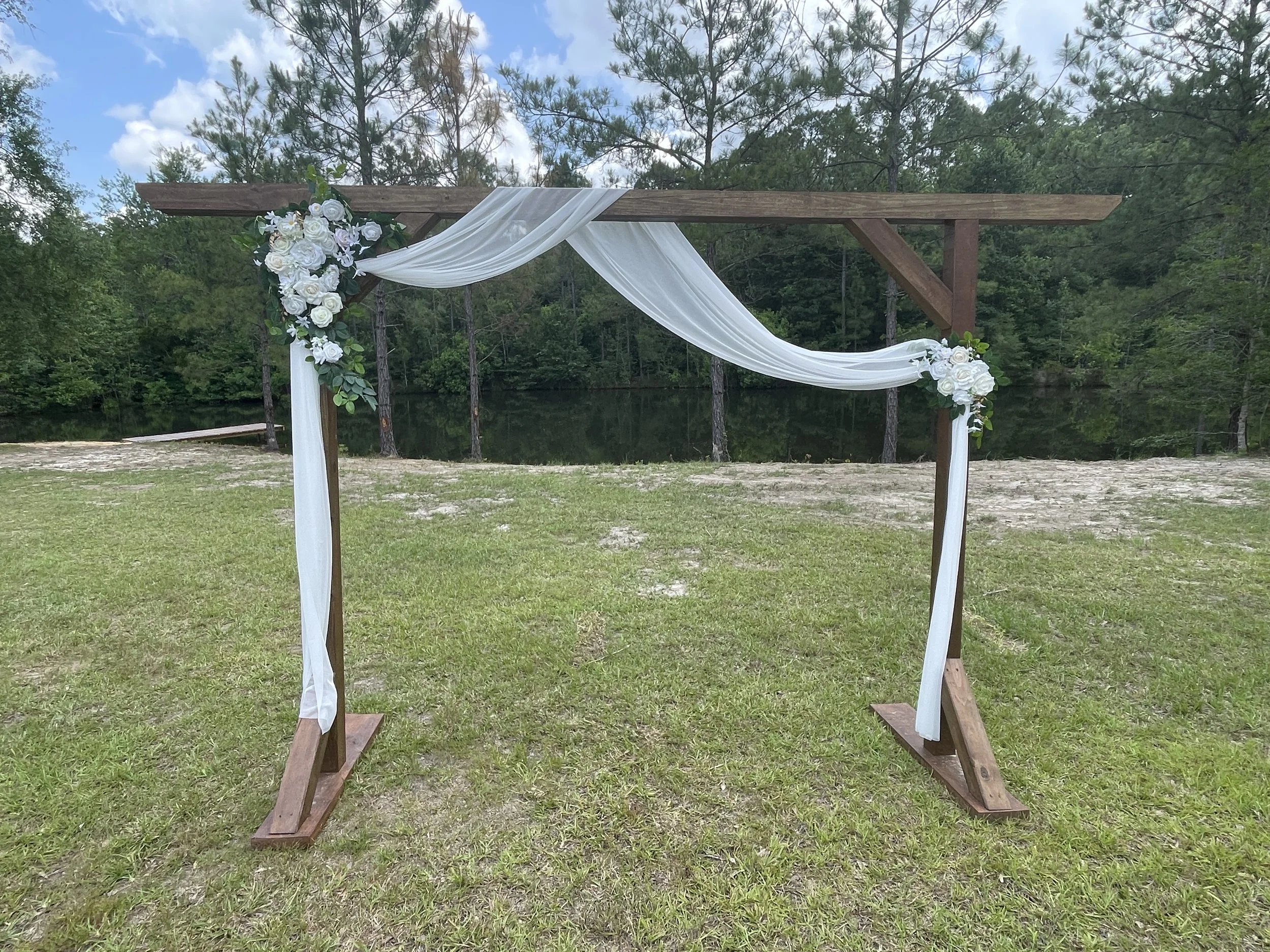 A wedding arch made of wooden beams decorated with white flowers, greenery, and flowing white fabric, set outdoors near a lake with trees in the background.