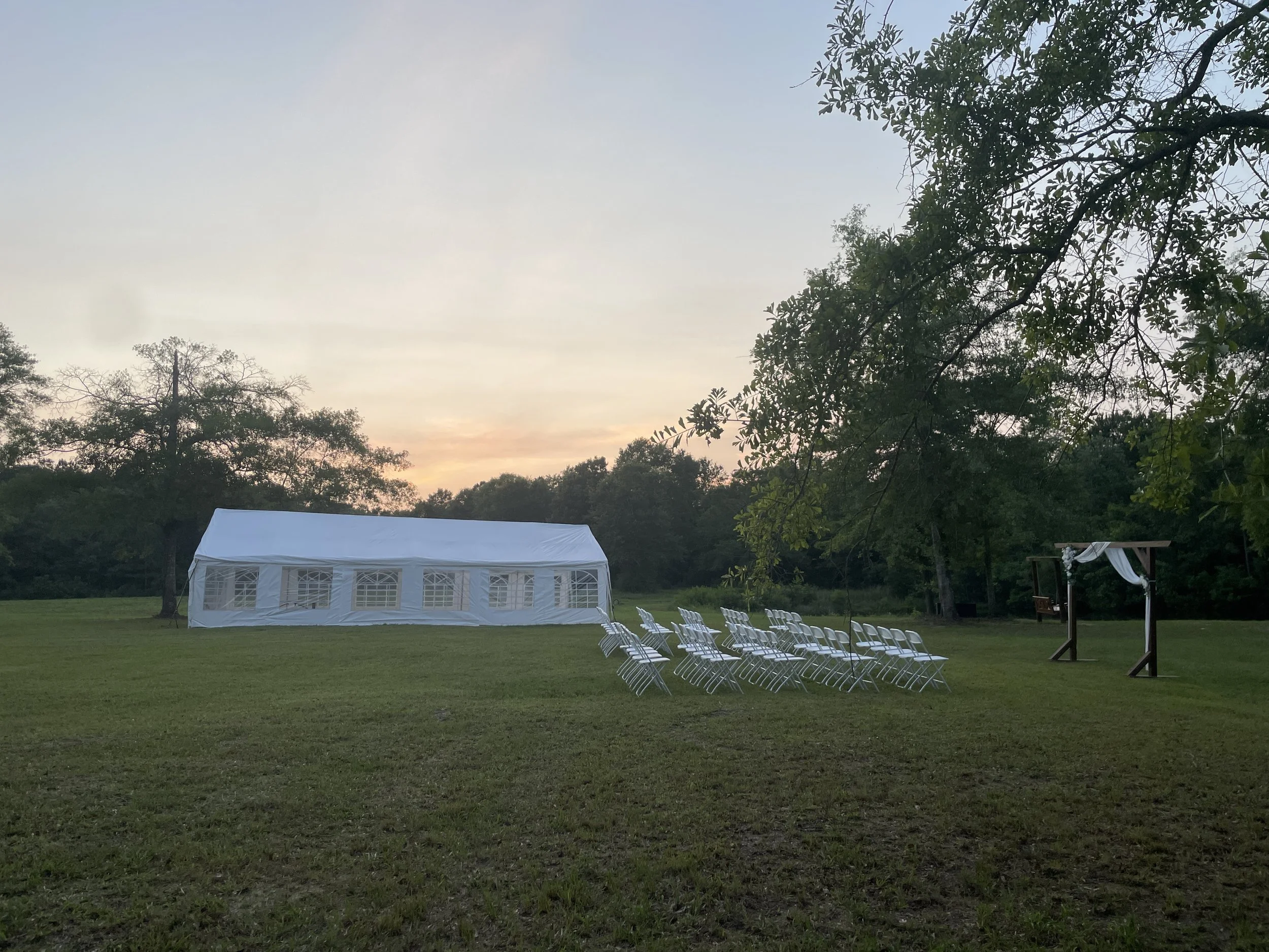 An outdoor event setup with white chairs arranged on a grassy field, a white tent at the center, and trees in the background with a sunset sky.