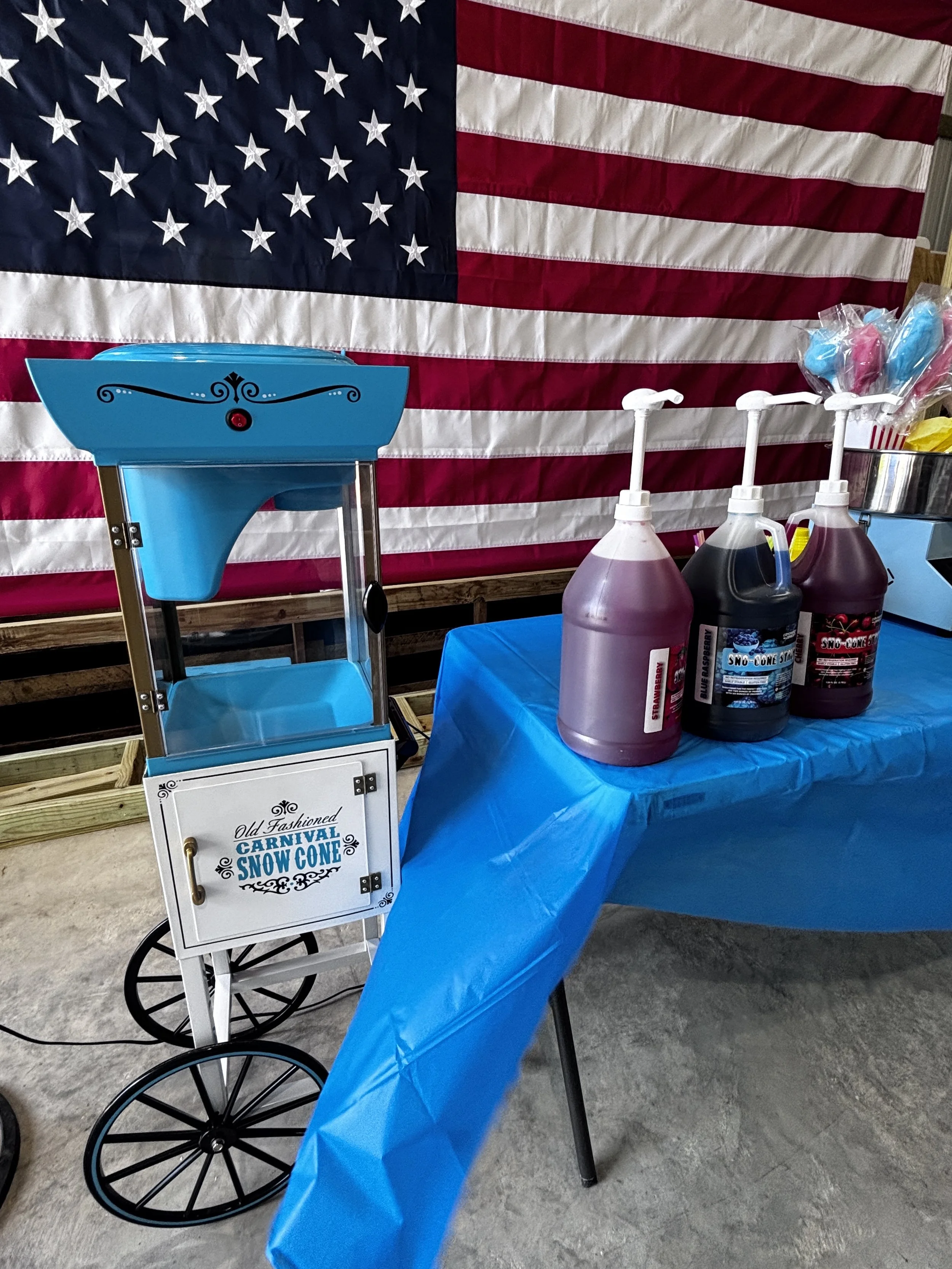 Carnival snow cone stand with a large American flag backdrop, featuring various syrups and a blue snow cone machine.