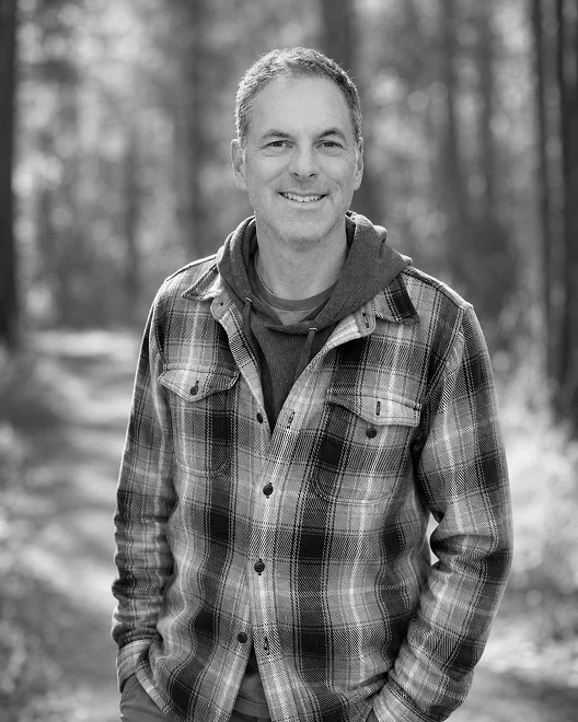 A smiling man outdoors in a forest, wearing a plaid jacket and a hoodie.