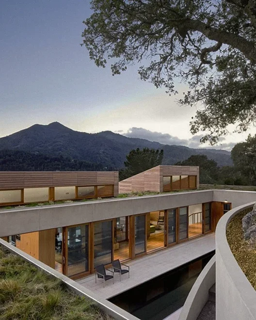 Modern house with large glass windows and wooden accents, integrated and built mountainous landscape at sunset.