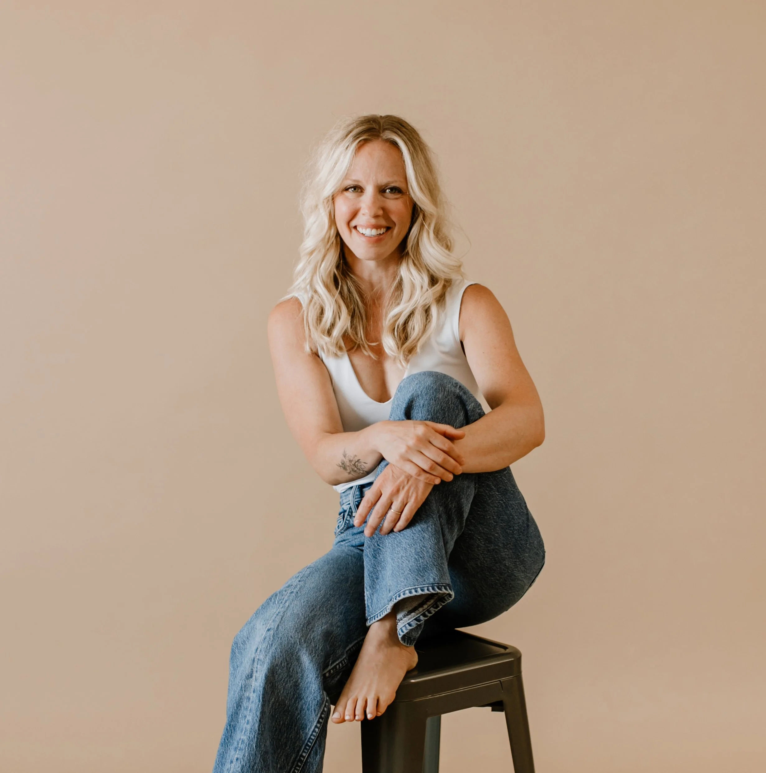 A woman with long, curly blonde hair, wearing a sleeveless white top, smiling. She is sitting on a black stool, with her arms resting on her crossed legs, one foot on the stool and the other arm resting on her knee. The background is plain beige.