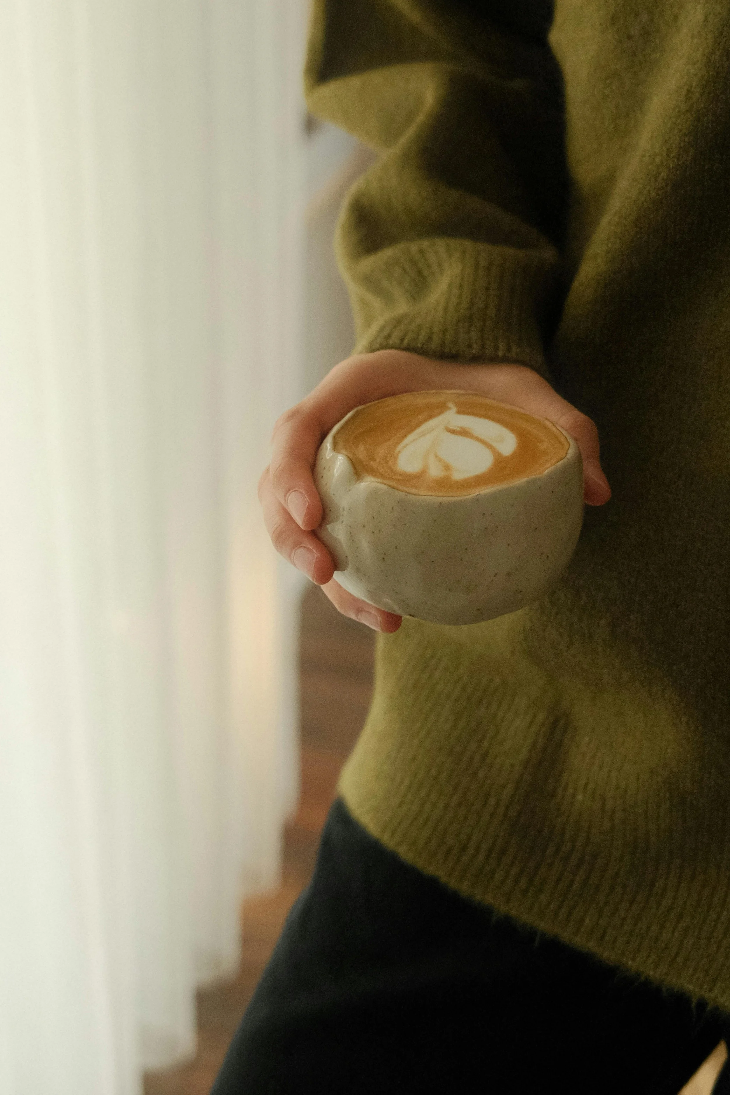 A person wearing a green sweater holding a cup of coffee or latte with foam art, standing near a window.