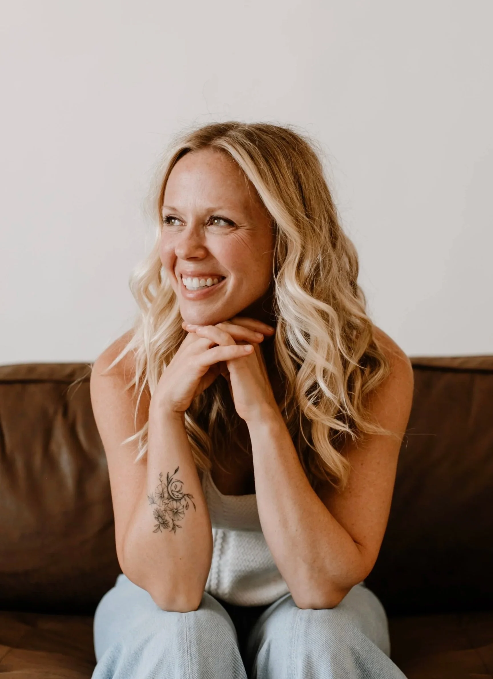 A woman with long wavy blonde hair sitting on a wooden bench with a tattoo of flowers on her left forearm, smiling and looking to her right, against a plain white wall.