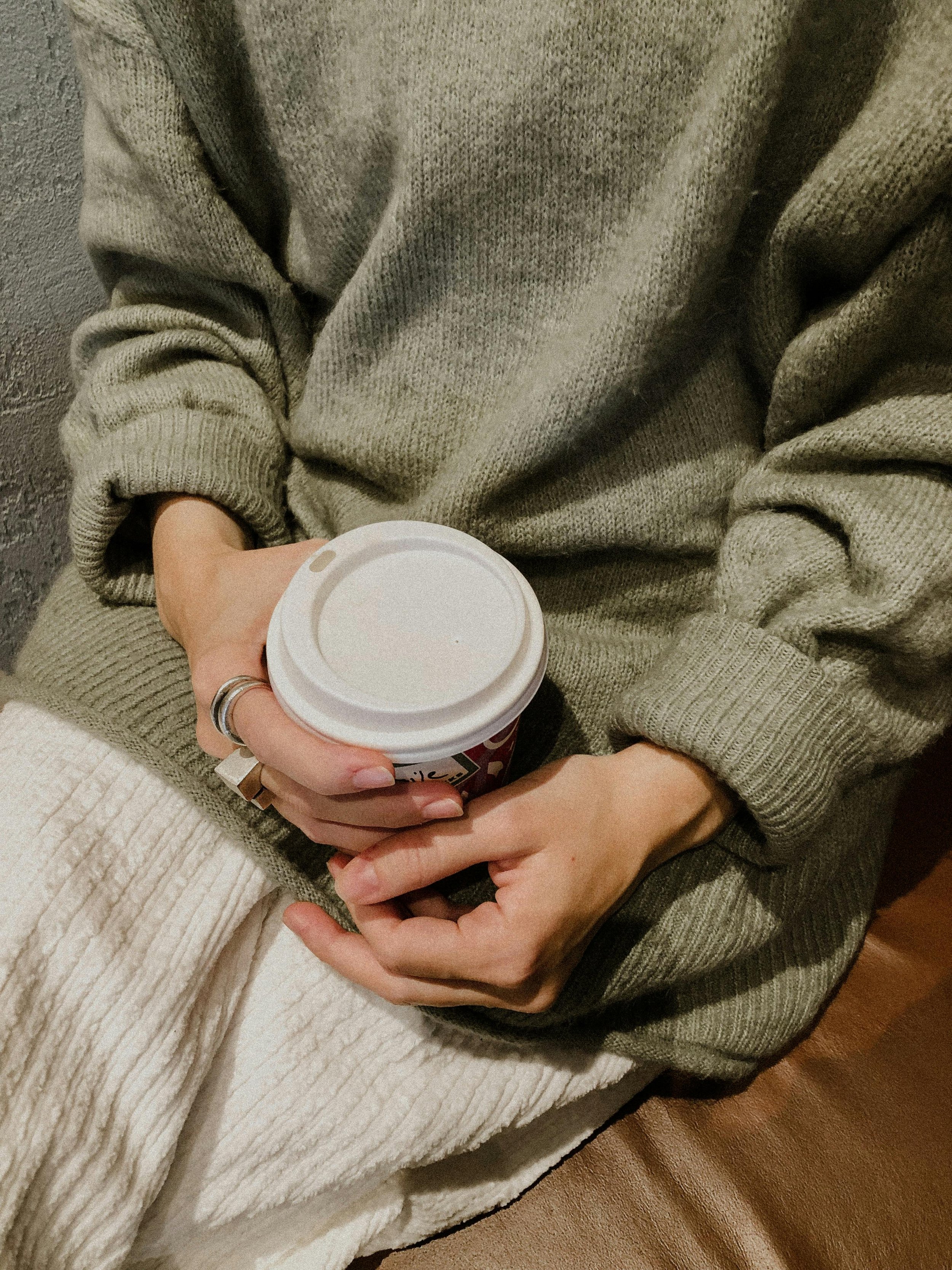 Person dressed in a cozy gray sweater holding a disposable coffee cup with a white lid.