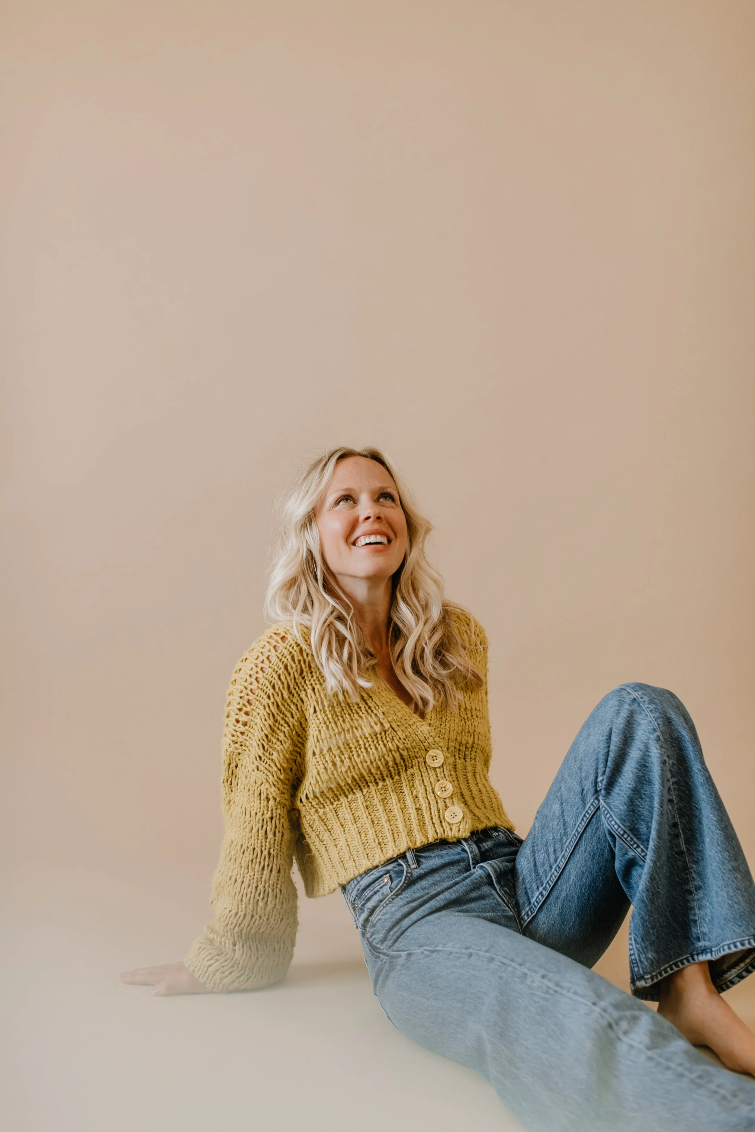 A young woman with long blonde hair, wearing a mustard yellow knitted sweater with buttons, sitting on the floor and smiling while looking up, against a plain beige background.