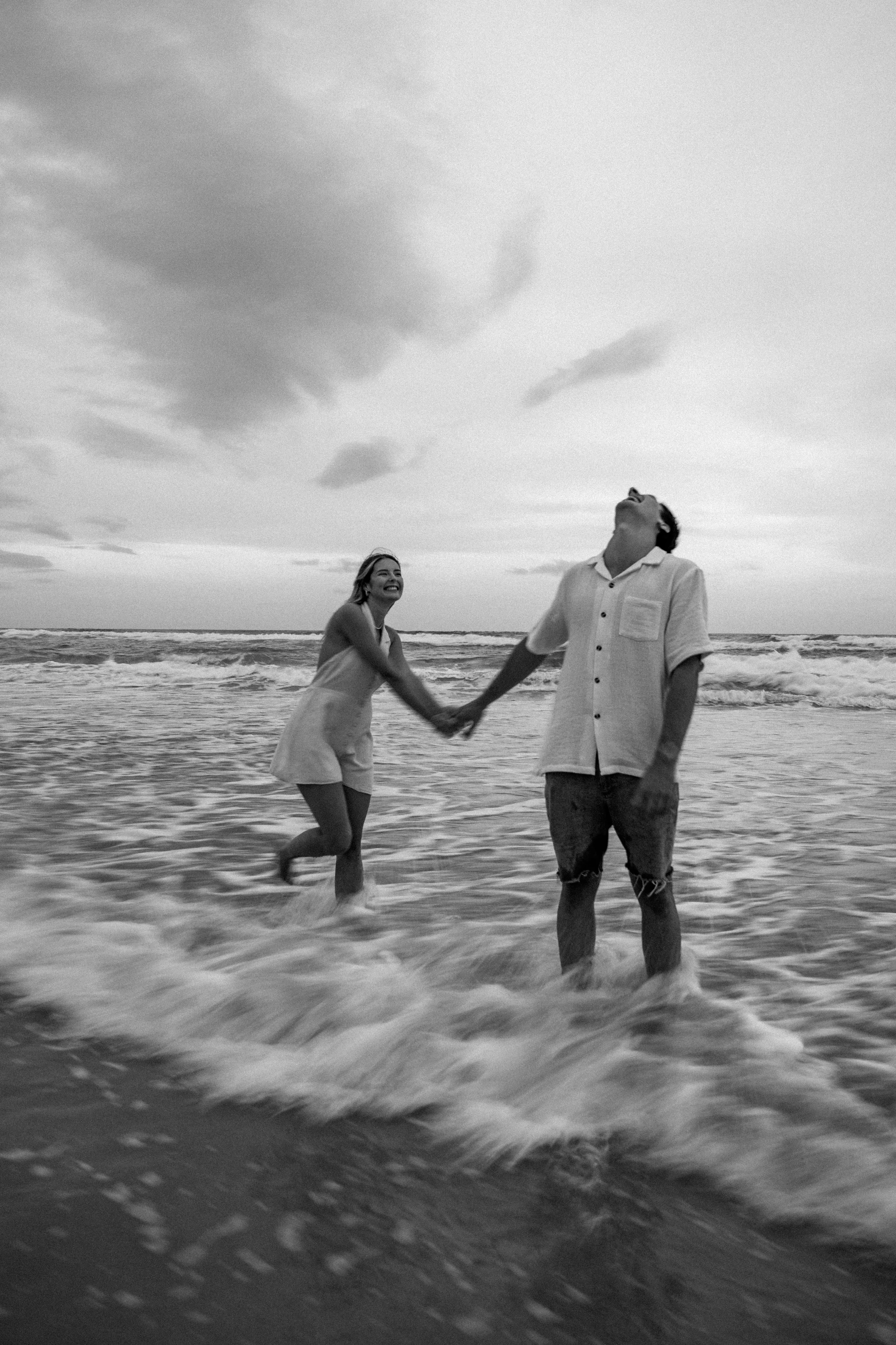 A black and white photo of a couple holding hands and playing in the ocean waves at the beach, with clouds in the sky.