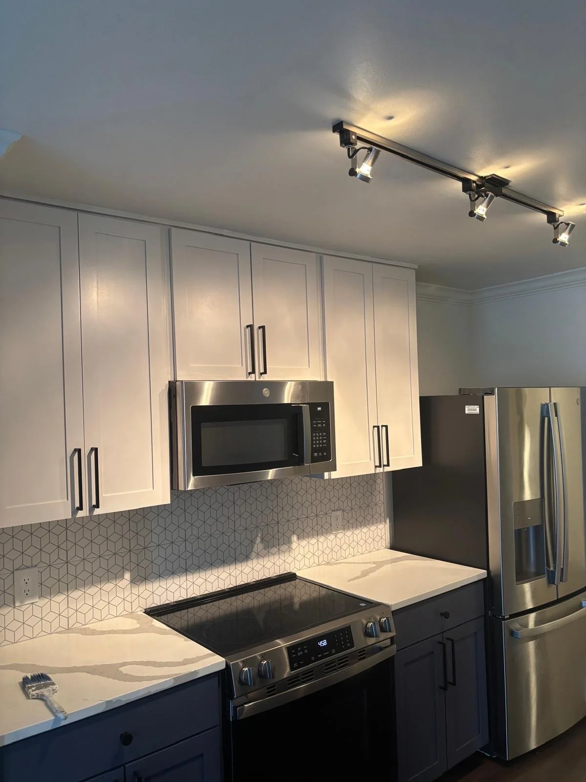 Modern kitchen with white cabinets, stainless steel microwave and refrigerator, black stovetop, white marble countertops, geometric tile backsplash, and ceiling track lighting.