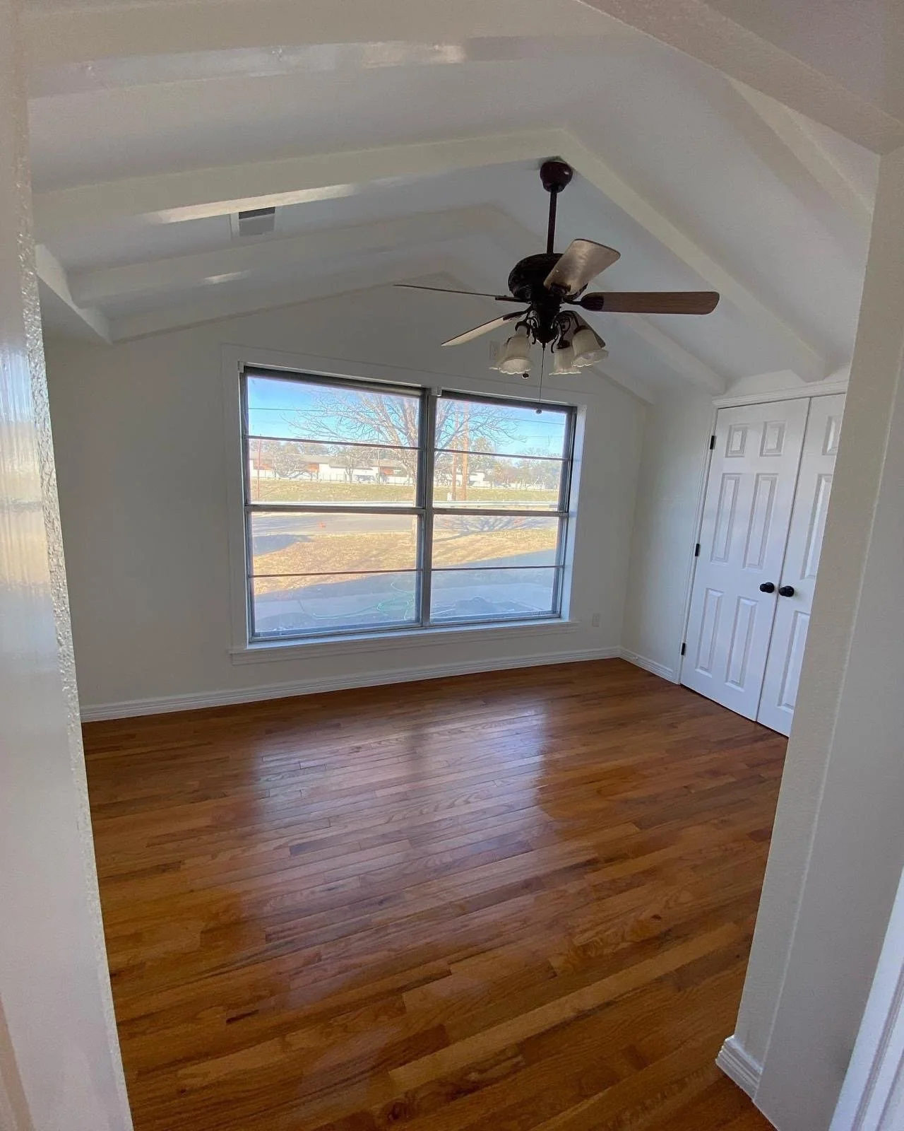 Empty room with hardwood flooring, white walls, and a large window showing an outdoor scene with trees and a street, ceiling fan with lights, and a closed closet door.