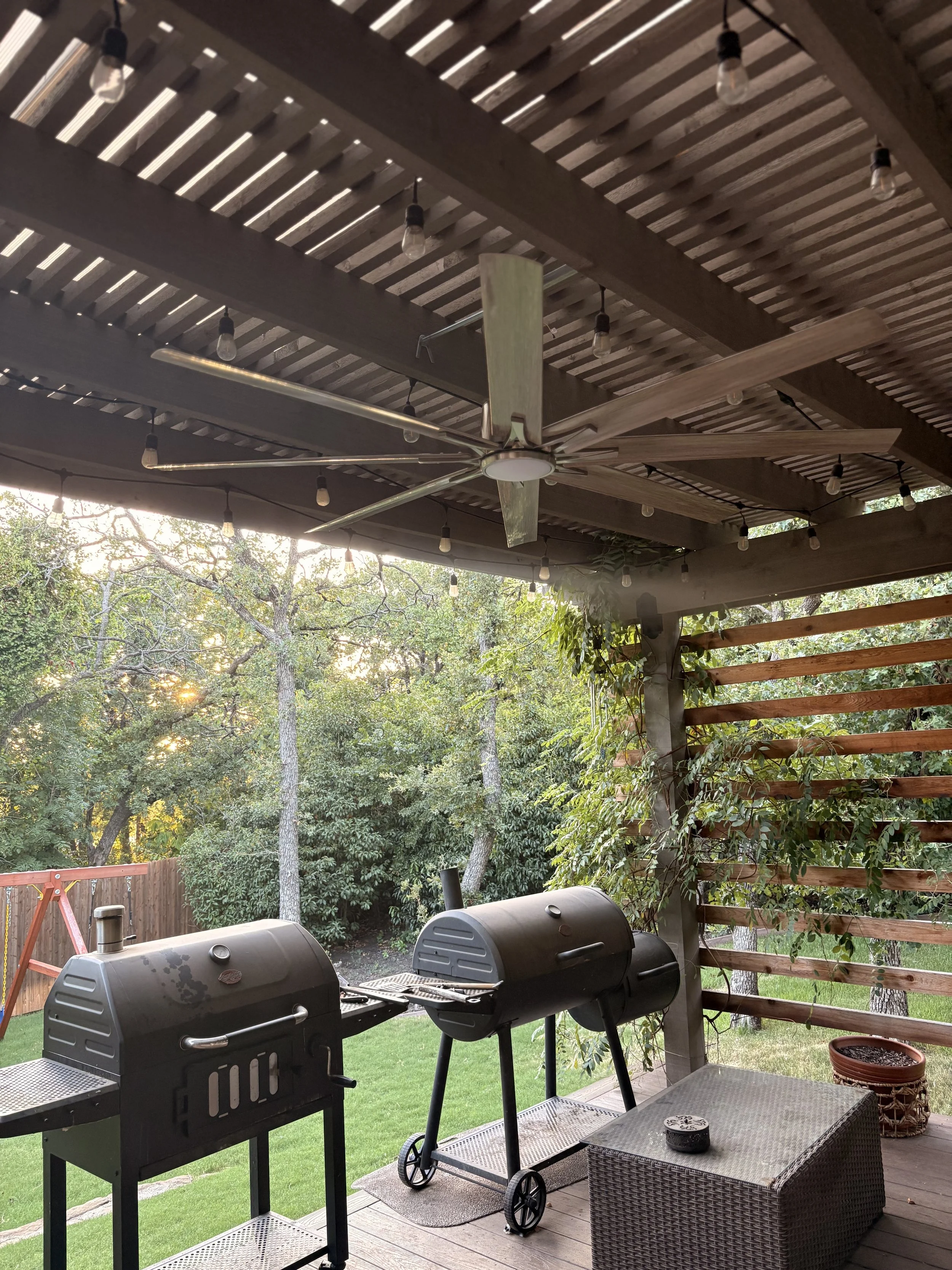 Backyard patio with two black grills, a patio table with a small decorative box, a woven basket, a potted plant, a wooden privacy fence, a pergola with string lights, and a ceiling fan, surrounded by trees and greenery.