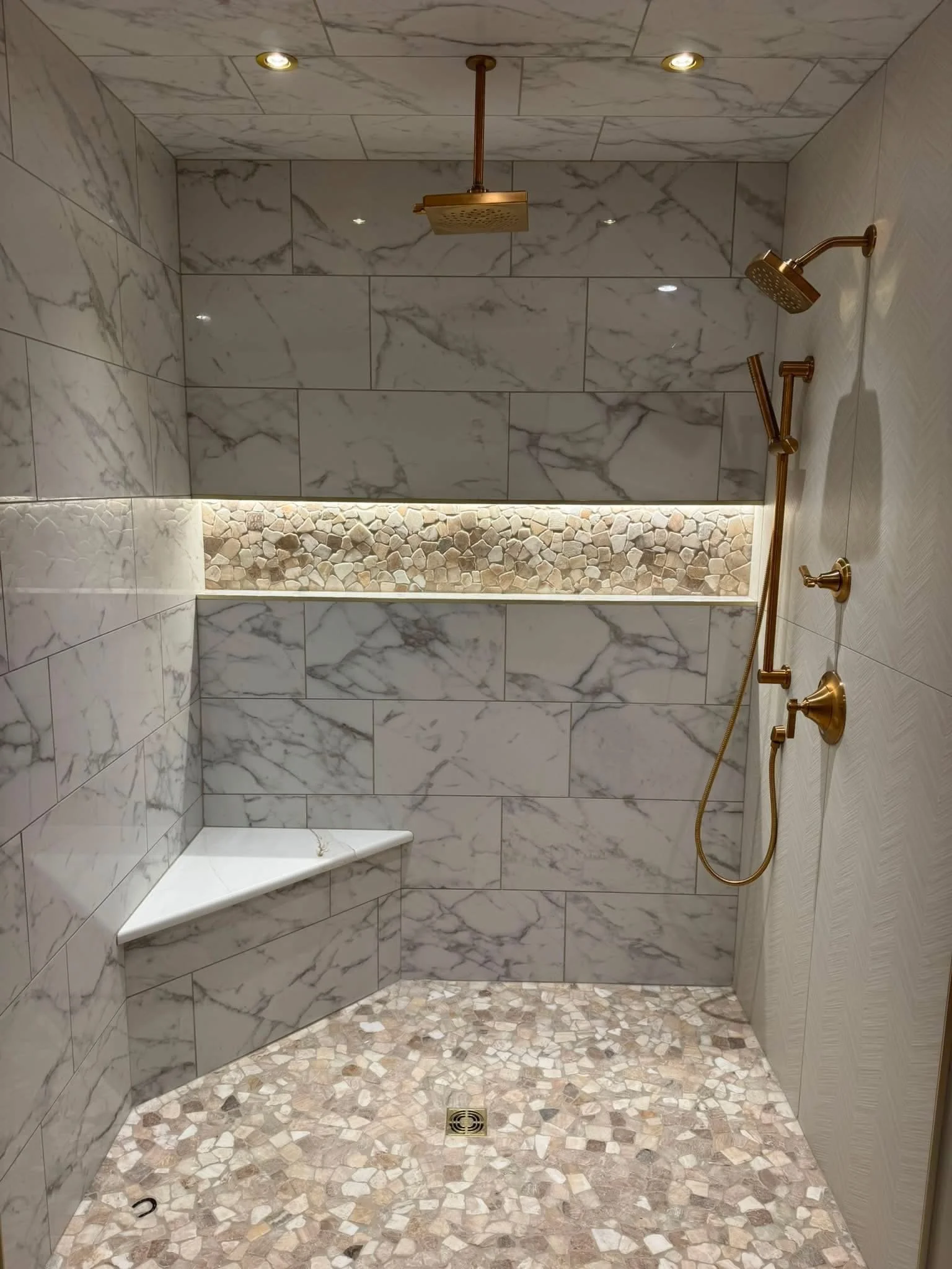 A walk-in shower with marble tiles on the walls and a decorative pebble tile pattern on the floor. It features a built-in corner bench, a rain showerhead, a handheld showerhead, and a wall-mounted shower arm, all finished in brushed gold. There are recessed ceiling lights and an LED-lit niche with small stones.