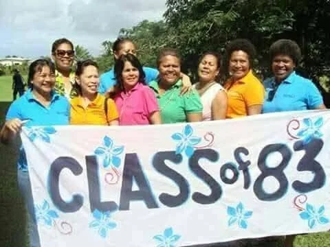 Group of women holding a banner that says 'Class of 83' with blue and pink floral decorations outdoors.
