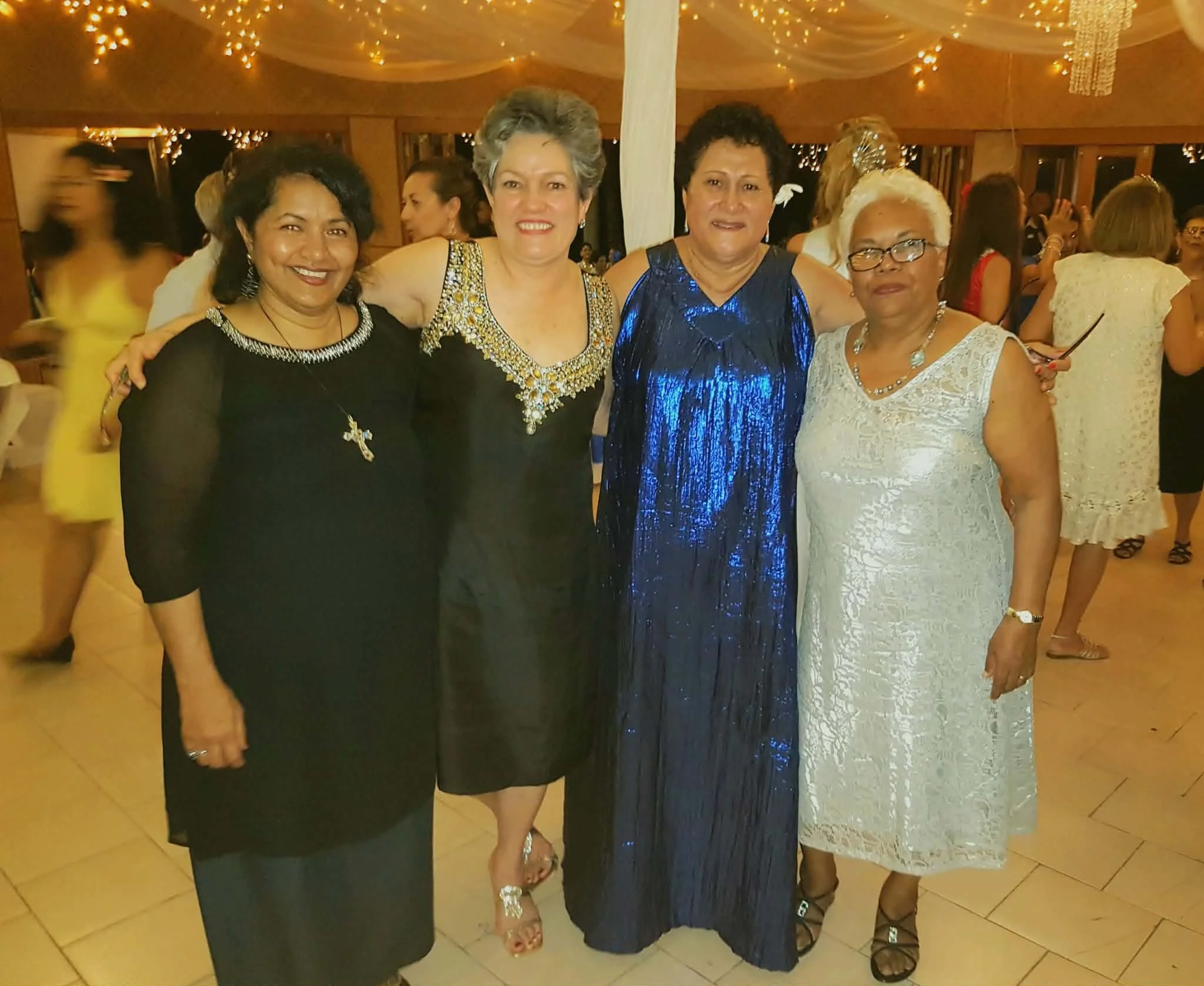 Four women standing close together at a festive event, dressed in elegant evening gowns, smiling for the camera, in a decorated venue with string lights and other attendees in the background.