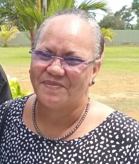 An older woman with short gray hair, wearing purple glasses, earrings, a beaded necklace, and a black and white patterned dress, standing outdoors with palm trees and a fence in the background.