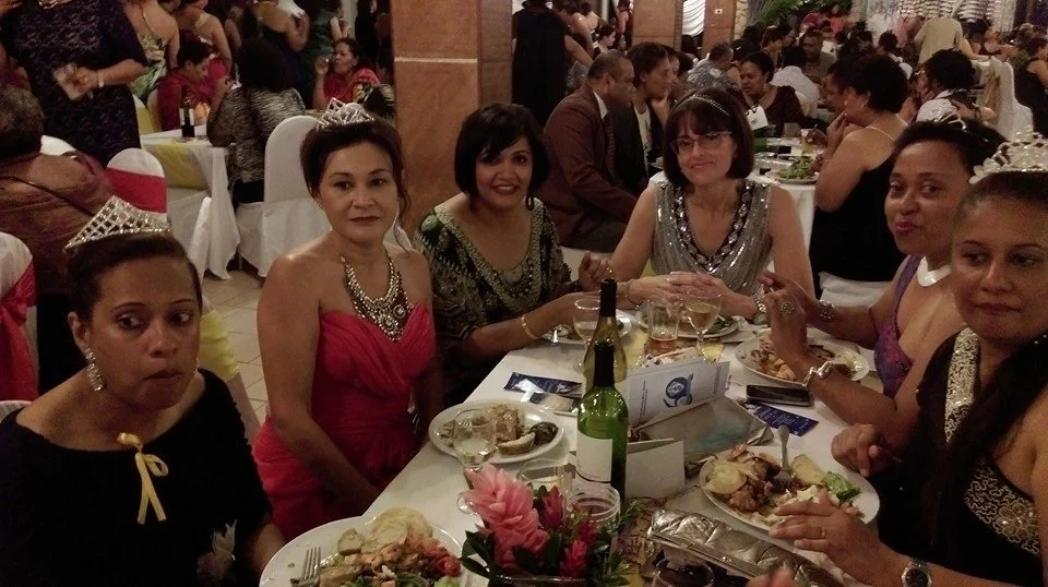 Group of women at a banquet table during a celebration or party, some wearing tiaras and dressed in elegant attire, with plates of food, drinks, and floral centerpieces.