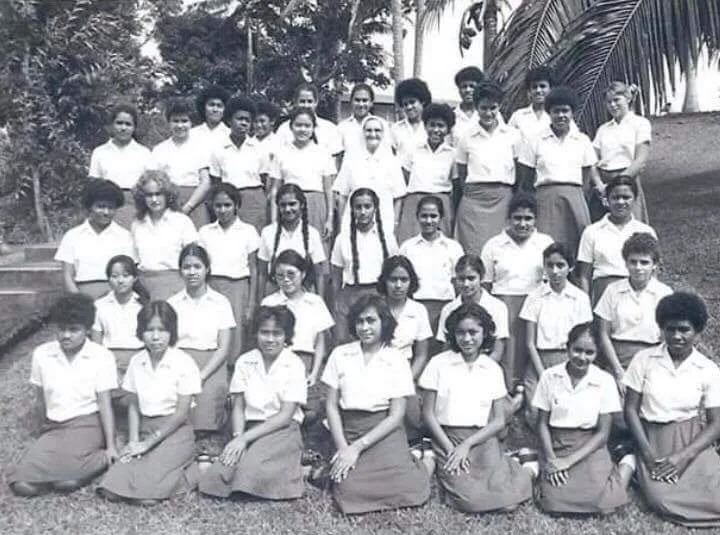 Group of students and teachers outdoors, posing for a photo. Students in white shirts and skirts, teachers in white shirts and skirts or pants. Trees in background.