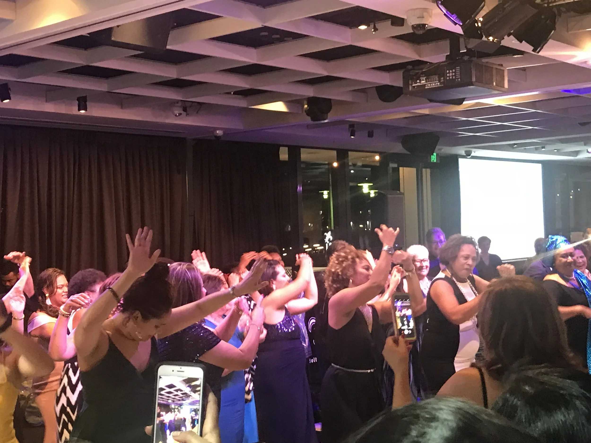 People in formal attire dancing and celebrating inside a banquet hall with purple and yellow lighting.