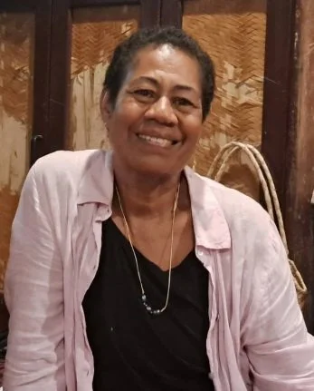 A smiling woman with short, curly hair wearing a light pink shirt over a black top, sitting indoors with wooden paneling and woven decor behind her.