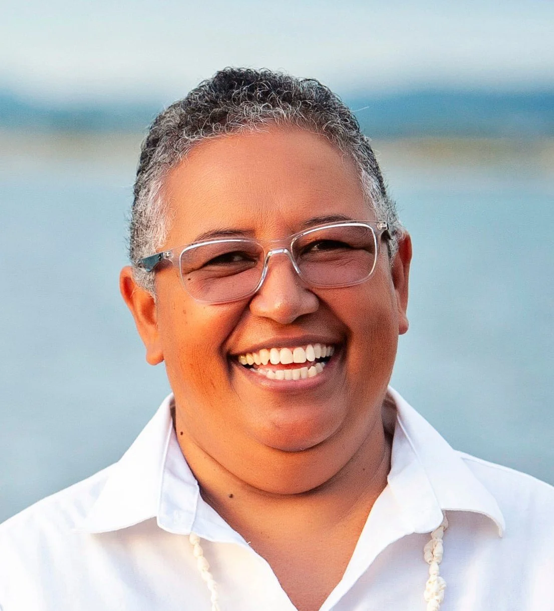 A smiling person with short gray hair, wearing glasses, a white shirt, and a necklace, standing outdoors near a body of water during sunset or sunrise.