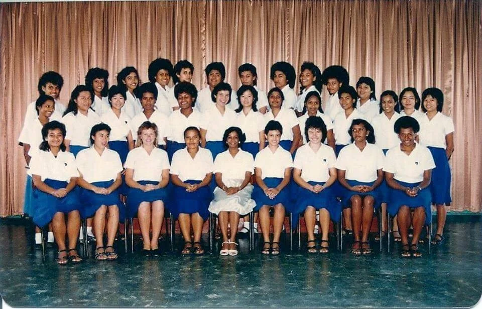 Group of young women in school uniforms posing for a class photo on stage with a pink curtain background.