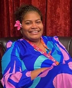 Smiling woman with dark hair in a colorful blue and pink dress, pink flower in her hair, sitting on a black chair with a red curtain background.