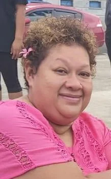 Close-up of a woman wearing a pink top, smiling outdoors with a parked car in the background.