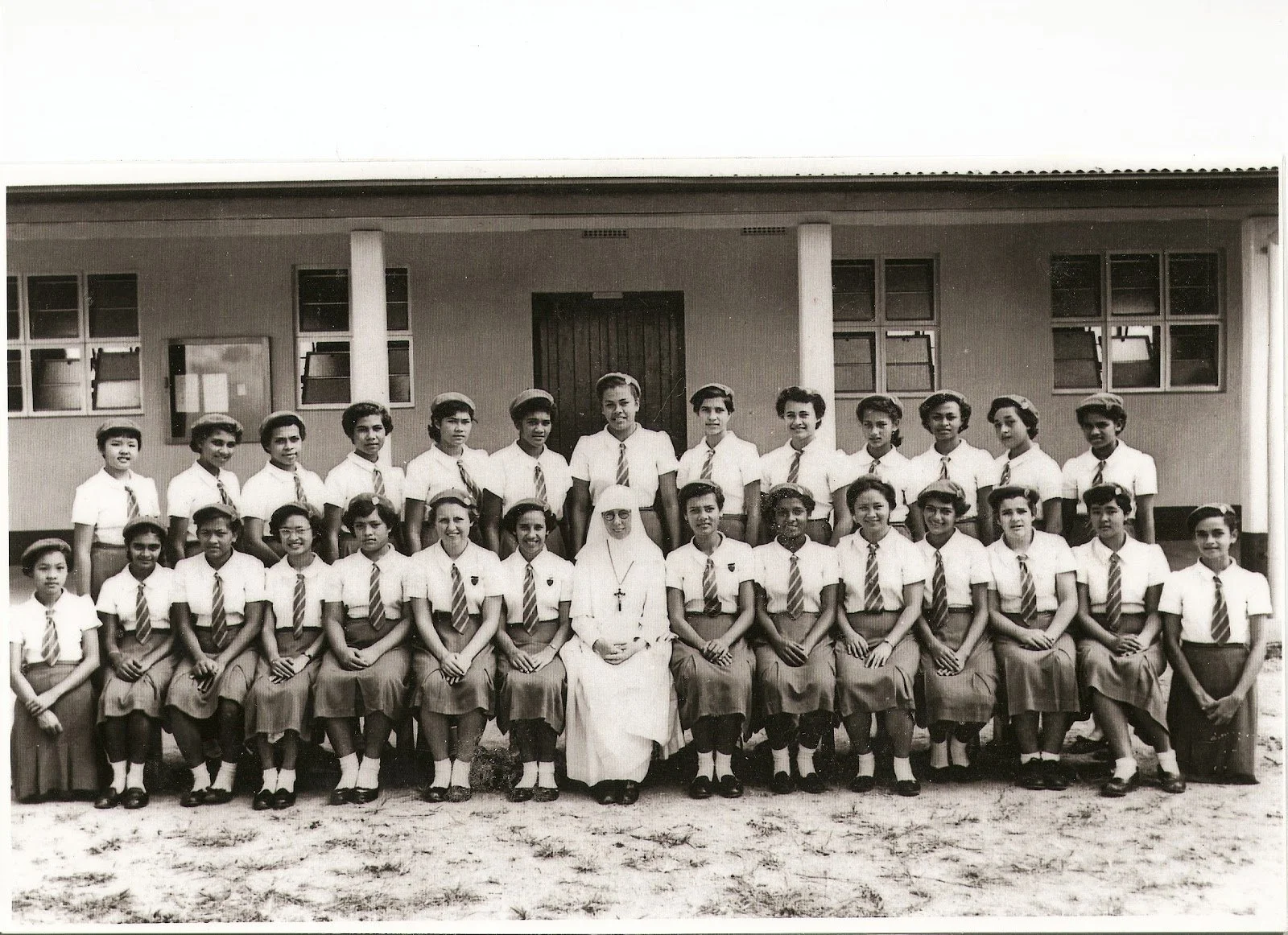 Black and white photograph of a group of school children and a nun, arranged in three rows in front of a school building, all dressed in uniforms with white shirts, striped ties, and skirts or shorts.