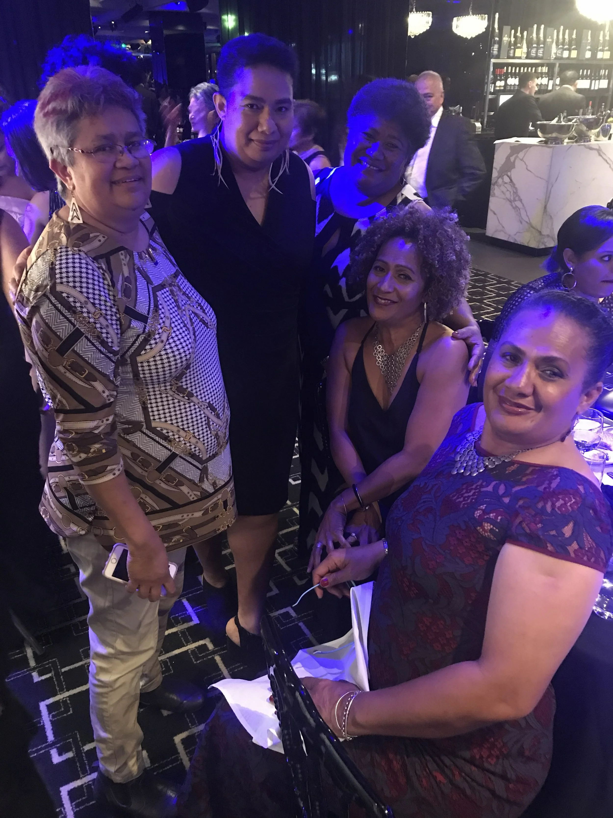 A group of women and a man posing together at a social event in a dimly lit, elegant setting. The women are dressed in stylish outfits with jewelry, and the man is dressed in a dark outfit. The background shows a bar area with bottles and glasses, an