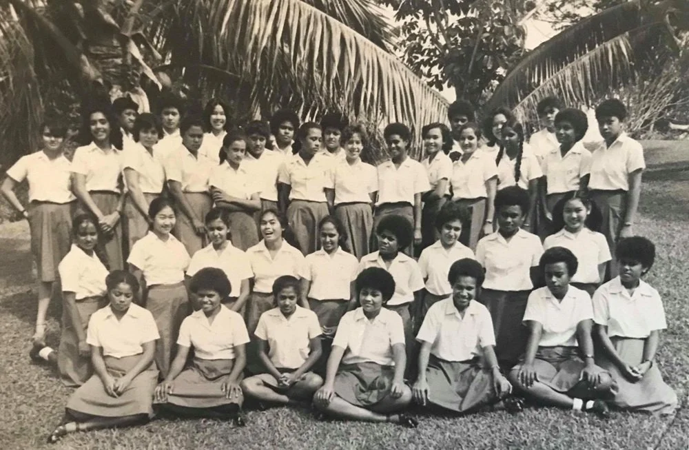 Black and white photo of a school group of girls and boys in uniform, standing outdoors on grass with large palm trees in the background.