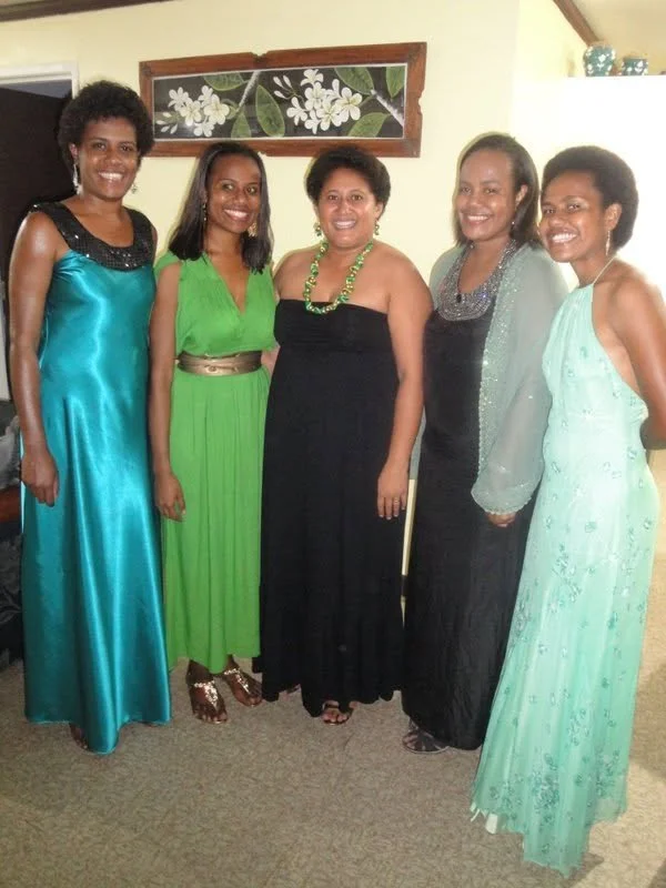 Five women in colorful evening dresses standing indoors, smiling at the camera.