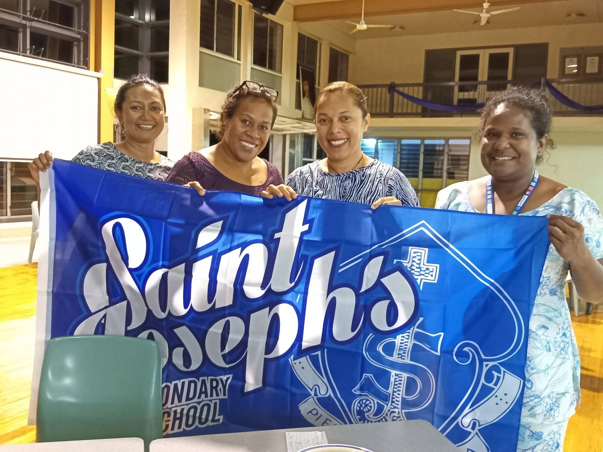 Four women standing together indoors, holding a blue banner with white text, smiling at the camera.