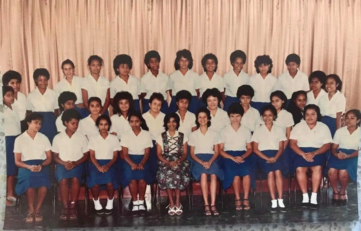 Group photo of school girls and teacher in front of curtains, all wearing white shirts and blue skirts.